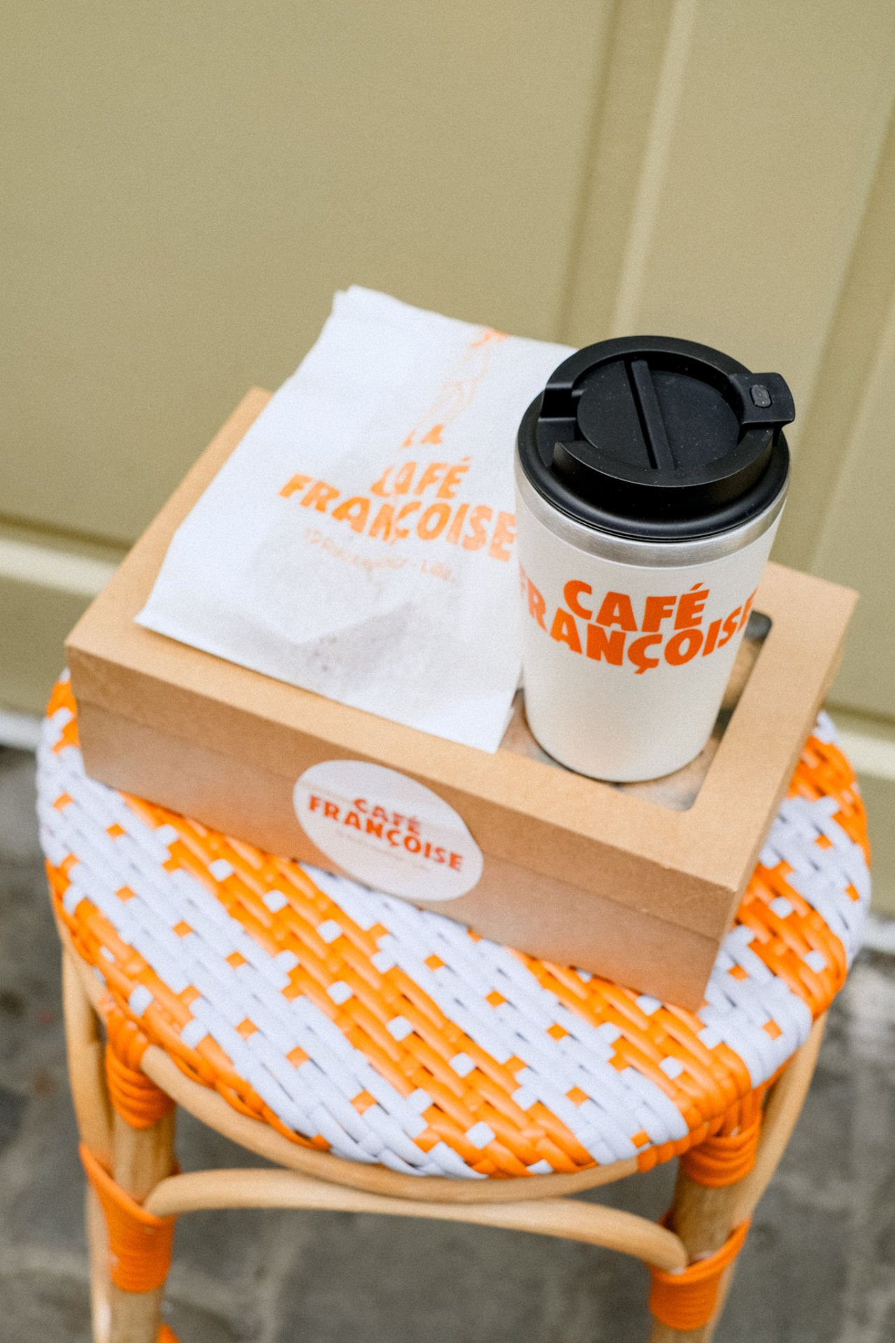 Gobelet et sachet Café Françoise à emporter posés sur un tabouret en rotin orange